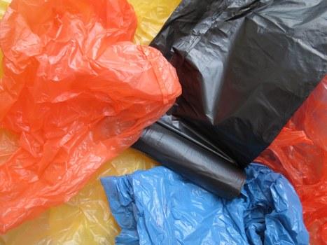 Close-up image displaying a collection of brightly coloured plastic rubbish bags, including orange, black, blue, and yellow, crumpled and piled together on what appears to be an outdoor surface. The bags are made of thin, shiny plastic material with a slightly wrinkled texture, and the black bag is partially overlapping the blue one, with the orange bag prominently shown in the foreground. The environment suggests a setting suitable for waste collection, possibly a driveway or pavement, with no other objects or background elements visible. The image visually supports rubbish removal services in Chelsea, highlighting the typical collection of waste bags prepared for disposal or removal by companies such as Business Waste Removal Chelsea.
