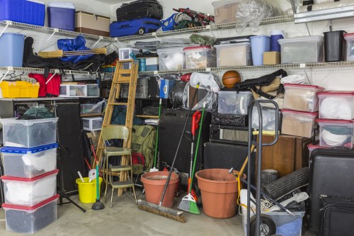A cluttered storage area with metal shelving units filled with various plastic storage boxes, containers, and bags of different sizes and colours, mainly transparent, blue, and black, stacked and arranged haphazardly. Some containers have lids, while others are open or partially covered. In the foreground, there are several large terracotta plant pots, a small yellow mop bucket with a mop leaning against it, and yard tools such as a broom and rake. A wooden step ladder and a couple of folding chairs are positioned amongst the items, along with a wheeled hand truck and a partially visible black suitcase. The scene appears to be indoors or in a garage, with a concrete floor and a white wall in the background. The area looks used for storage of household, gardening, and miscellaneous items, and it is characteristic of a typical rubbish accumulation or items to be disposed of by a waste removal service such as Business Waste Removal Chelsea operating in the Chelsea area.