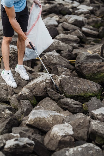A person wearing white sneakers, black shorts, and a blue t-shirt is standing on a rocky shoreline, using a grabber tool to pick up debris or small waste items among large, irregularly shaped stones w