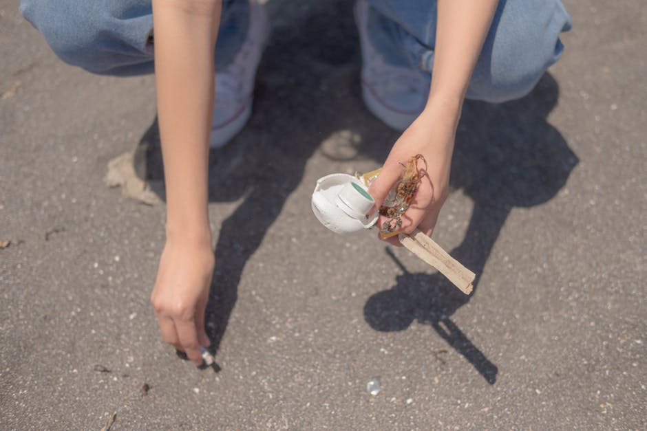 A person wearing white sneakers, black shorts, and a blue t-shirt is standing on a rocky shoreline, using a grabber tool to pick up debris or small waste items among large, irregularly shaped stones w