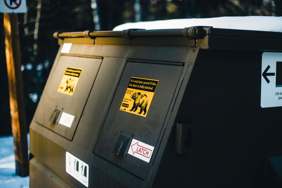 A large grey outdoor waste disposal container with a sloped lid, situated on a paved surface, likely in a residential or commercial area. The container features yellow and black warning stickers with