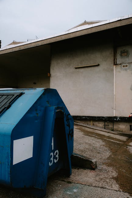 A large grey outdoor waste disposal container with a sloped lid, situated on a paved surface, likely in a residential or commercial area. The container features yellow and black warning stickers with